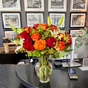 Evening Ember arrangement with red and orange roses, ivory lilies, golden accents, and greenery in a clear glass vase by a Santa Monica florist