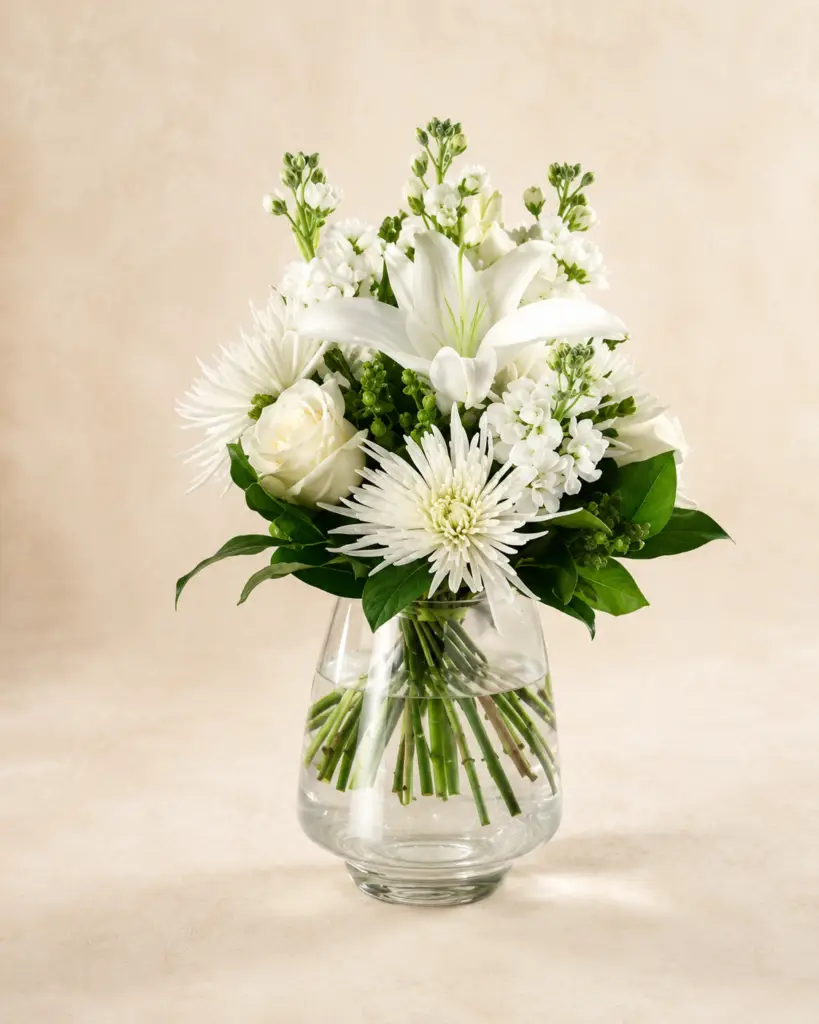 Ivory Bloom white flower arrangement with white lilies, roses, spider mums, seasonal accents, and greenery in a clear glass vase by a Santa Monica flower studio.