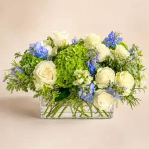 Ocean Meadow floral centerpiece with white roses, green hydrangea, soft blue blooms, white seasonal flowers, variegated greenery, and textural greens in a low glass vase by a Santa Monica florist.